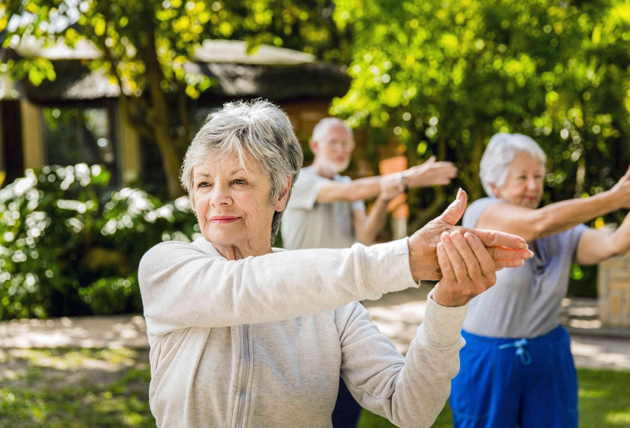 A group of elderly adults stand together outside on a sunny day and stretch during an exercise class.
